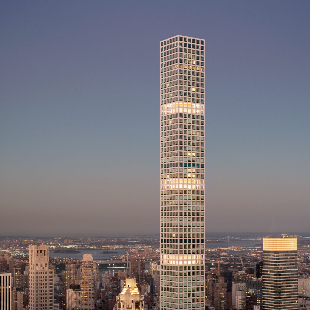 A tall, slender skyscraper with a grid-like facade stands prominently in the foreground against a city skyline at dusk. The sky is clear, and other buildings and a distant bridge are visible in the background.