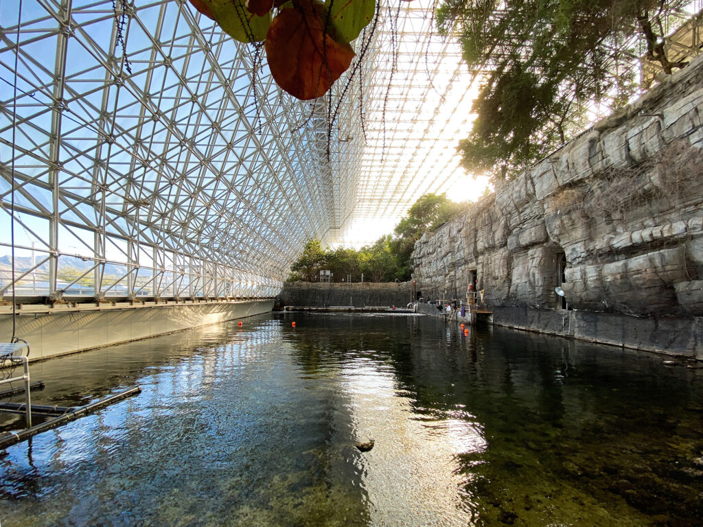 A geometric metal and glass structure arches over a tranquil pool surrounded by rocky walls and lush greenery. Sunlight filters through the framework, casting reflections on the waters surface. Orange buoys float near one edge.