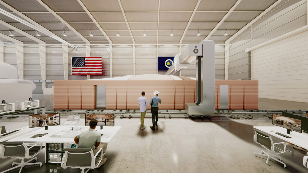 A large 3D printer is constructing a building inside a spacious warehouse. Two people stand observing the process. Tables and chairs are seen in the foreground. An American flag and another flag hang on the wall.