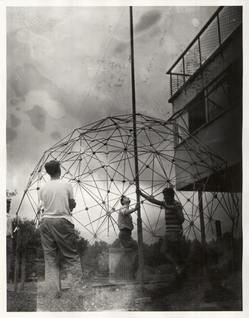 Black and white image of three individuals assembling a geodesic dome with a metal framework. One person is holding a pole, while the others assist. A building is partially visible on the right side. Overcast sky in the background.